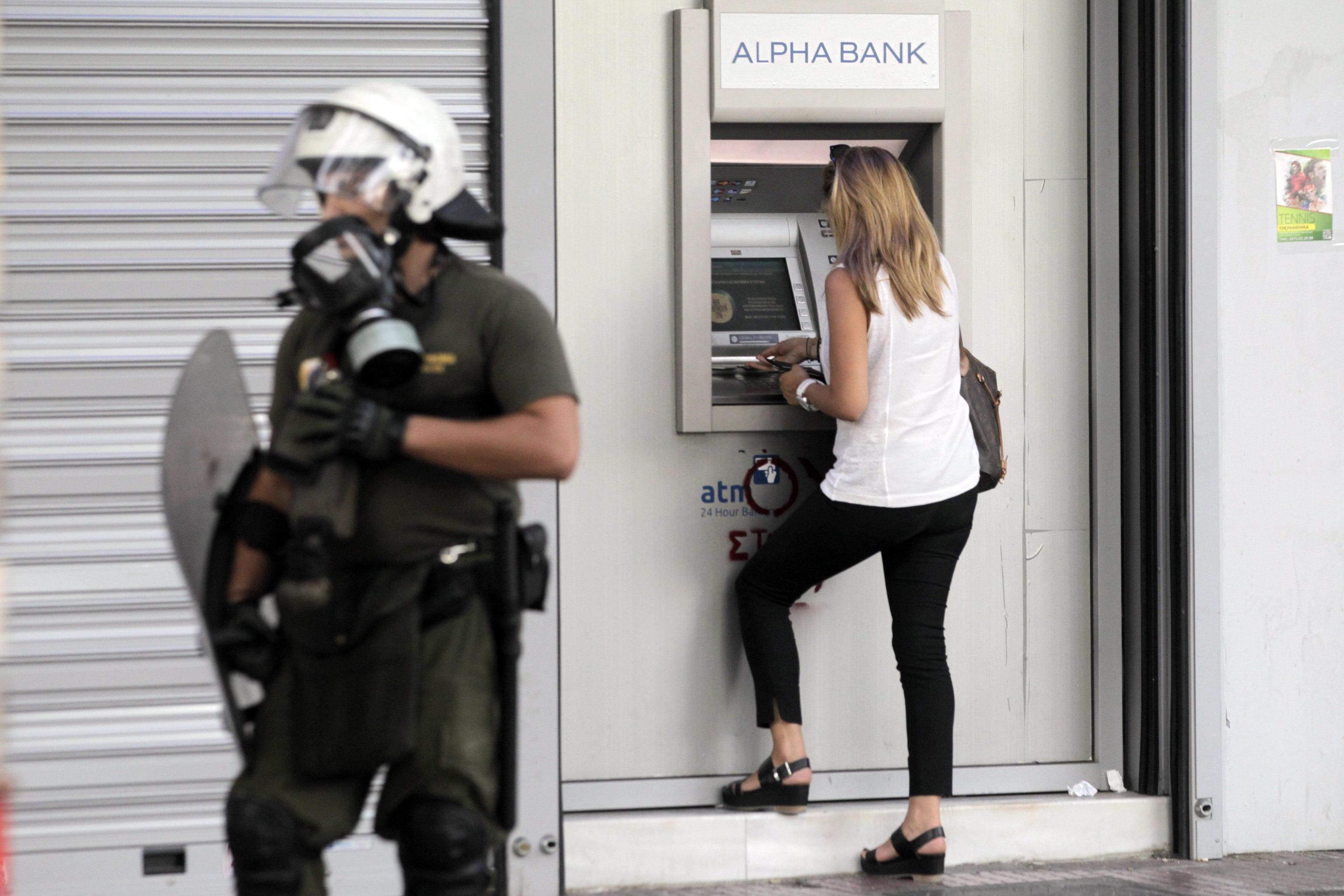 A woman withdraws cash from an ATM  in Athens while a riot policeman stands on duty
